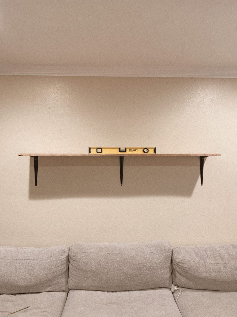 Wood shelf above couch, resting on brackets. A leveler is on top of the shelf. 