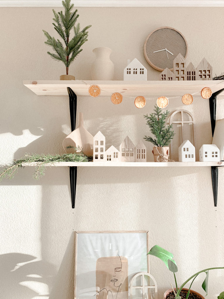 Wide view of orange garland hanging from wood shelf, surrounded by other small Christmas trees and wood/ceramic houses decor.