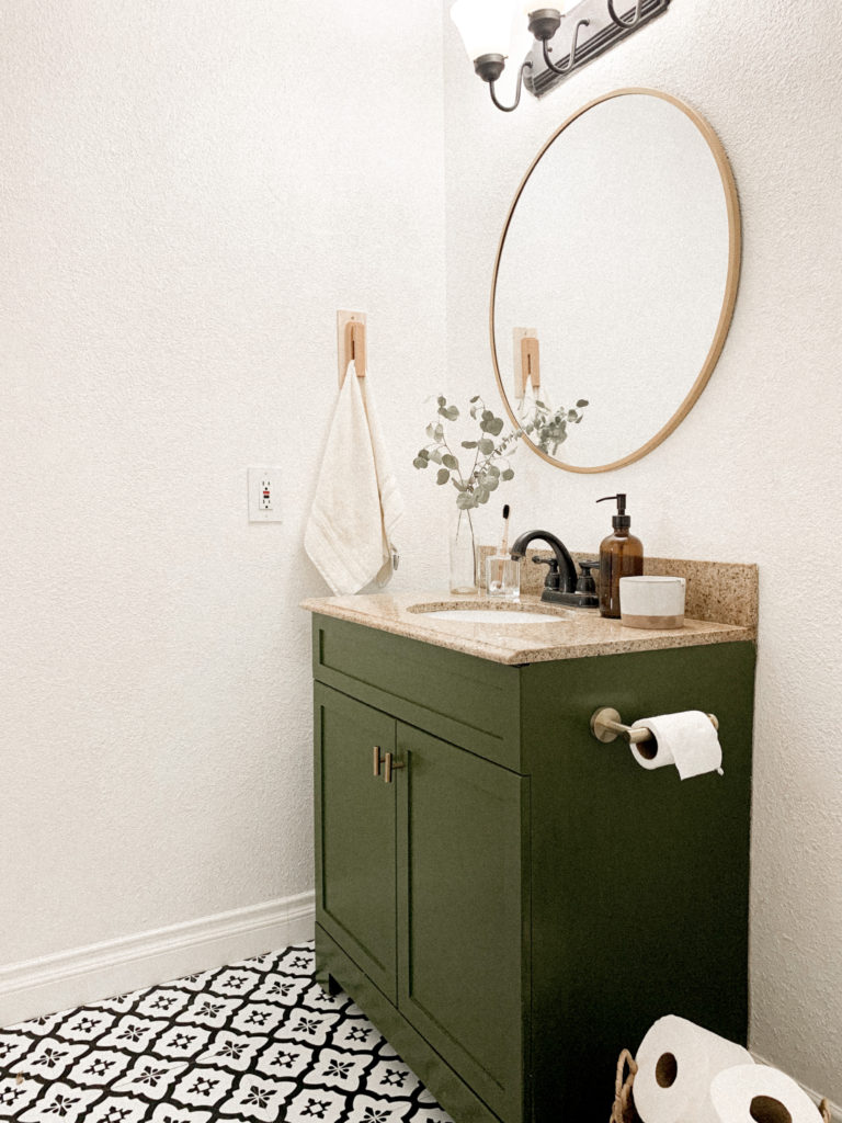 Medium view/side angle of dark green bathroom vanity, brass accents, and white and black stick and peel flooring.