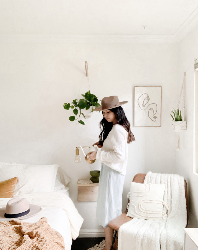 Girl in bohemian-style bedroom with boho hat, standing near bed and holding watering can below hanging planter.
